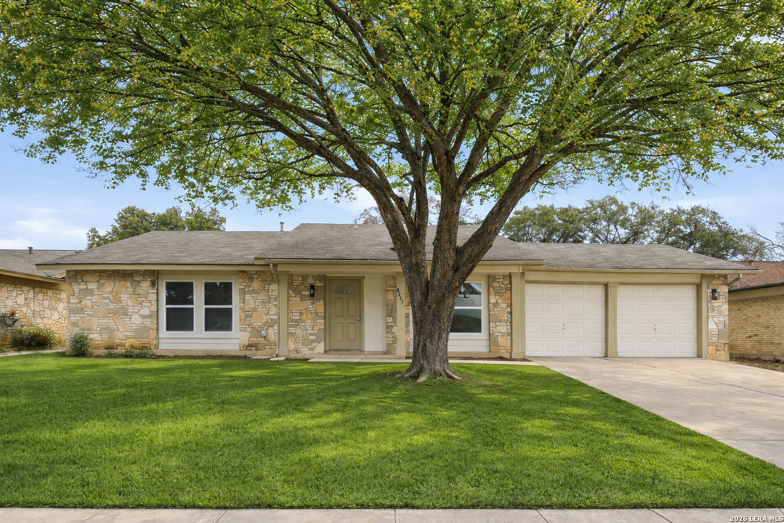 Front exterior of 8247 Lewiston St — stone ranch home with lush green lawn, large oak tree, and 2-car garage in spring